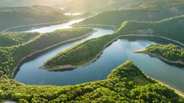 Autumn Short Break in the National Park | Gemünder Ferienpark Aerial view of a winding river surrounded by lush green forests and hills in sunlight