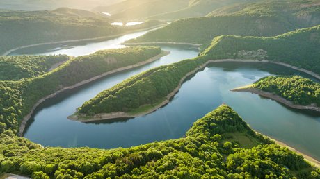 Ferien in der Eifel: ein erster Einblick Luftaufnahme eines mäandrierenden Flusses umgeben von grünen Wäldern und Hügeln im Sonnenlicht