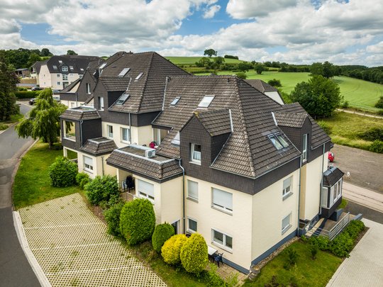 Wohnung mieten in Gemünd? Da haben wir was für Sie! Mehrfamilienhaus mit grauem Dach und grüner Landschaft bei bewölktem Himmel