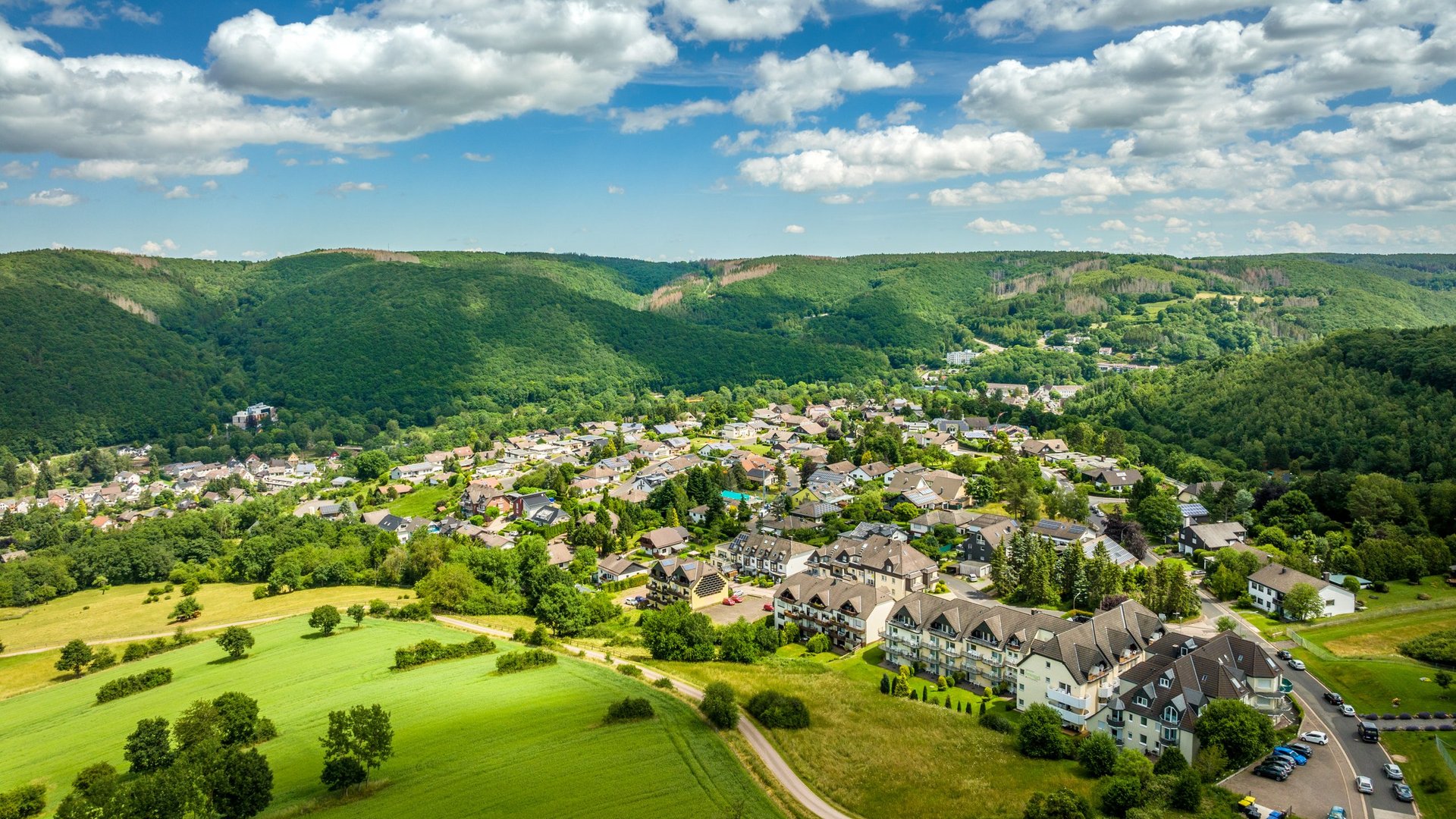 Urlaub in der Eifel mit Hund? Aber natürlich! Luftaufnahme eines Dorfes umgeben von grünen Wäldern und Feldern unter blauem Himmel