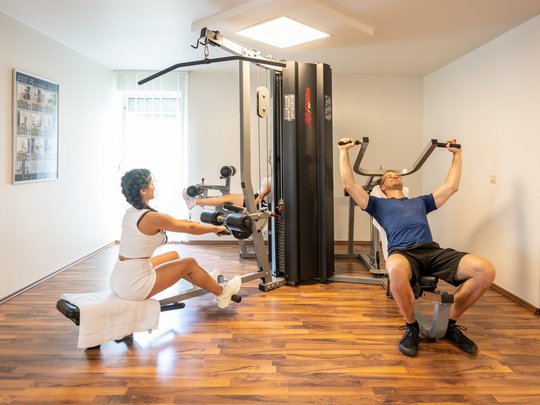 Wohnung mieten in Gemünd? Da haben wir was für Sie! Zwei Personen trainieren an Fitnessgeräten in einem hellen Raum mit Holzboden