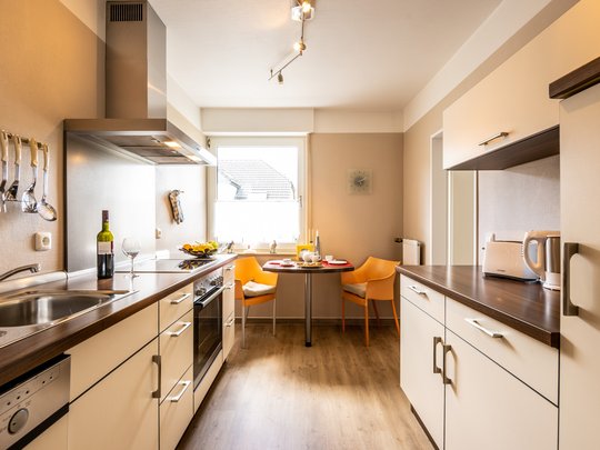 Ihre Unterkunft in Gemünd – Eifel-Urlaub voller Freiheit Moderne Küche mit Esstisch, zwei orangenen Stühlen, Weinflasche und Küchengeräten