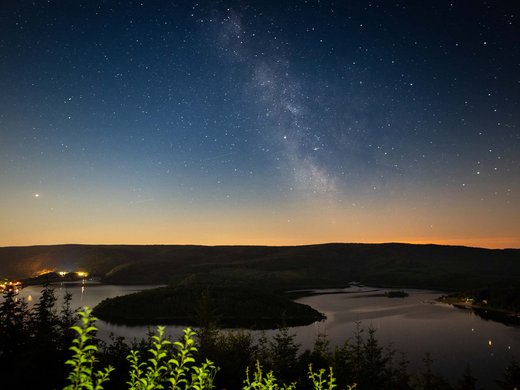 Sternenpark Eifel erleben – Urlaub unter dem Sternenhimmel Sternenhimmel mit Milchstraße über einem See und dunklen Hügeln bei Nacht