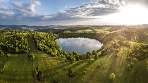 Ihre Wohnung in Gemünd in der Eifel: jetzt anfragen! Landschaft mit rundem See, grünen Wiesen und Sonnenstrahlen am Himmel