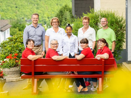 Wohnung mieten in Gemünd? Da haben wir was für Sie! Gruppe von neun lächelnden Personen, einige auf einer roten Bank sitzend, draußen im Grünen