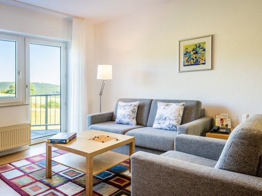 Ihre Unterkunft in Gemünd – Eifel-Urlaub voller Freiheit Helles Wohnzimmer mit grauem Sofa, Sessel, Holztisch und Blick ins Grüne