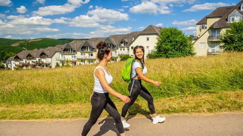 Ihre Wohnung in Gemünd in der Eifel: jetzt anfragen! Zwei Frauen wandern auf einem Weg mit Häusern und Wiesen im Hintergrund.