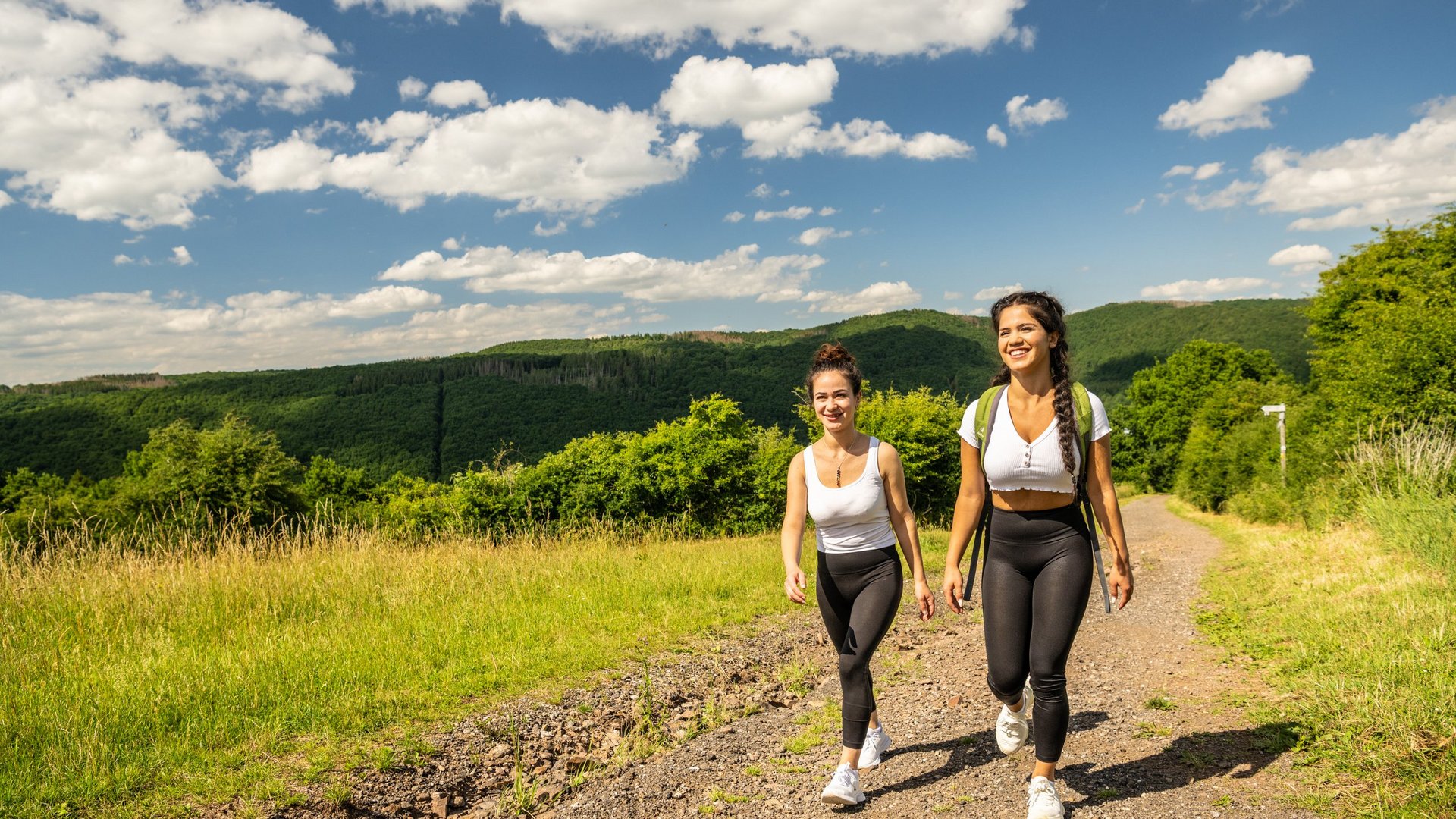 Eifel-Urlaub: Insidertipps aus Gemünd Zwei junge Frauen wandern auf einem Weg mit grünen Hügeln und blauem Himmel im Hintergrund