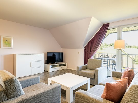 Ihre Unterkunft in Gemünd – Eifel-Urlaub voller Freiheit Helles Wohnzimmer mit grauen Möbeln, weißem Tisch, TV und Balkon mit Blick ins Grüne