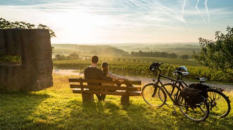 Ferien in der Eifel: ein erster Einblick Paar sitzt auf Bank mit Fahrrädern und blickt auf sonnige Weinberglandschaft