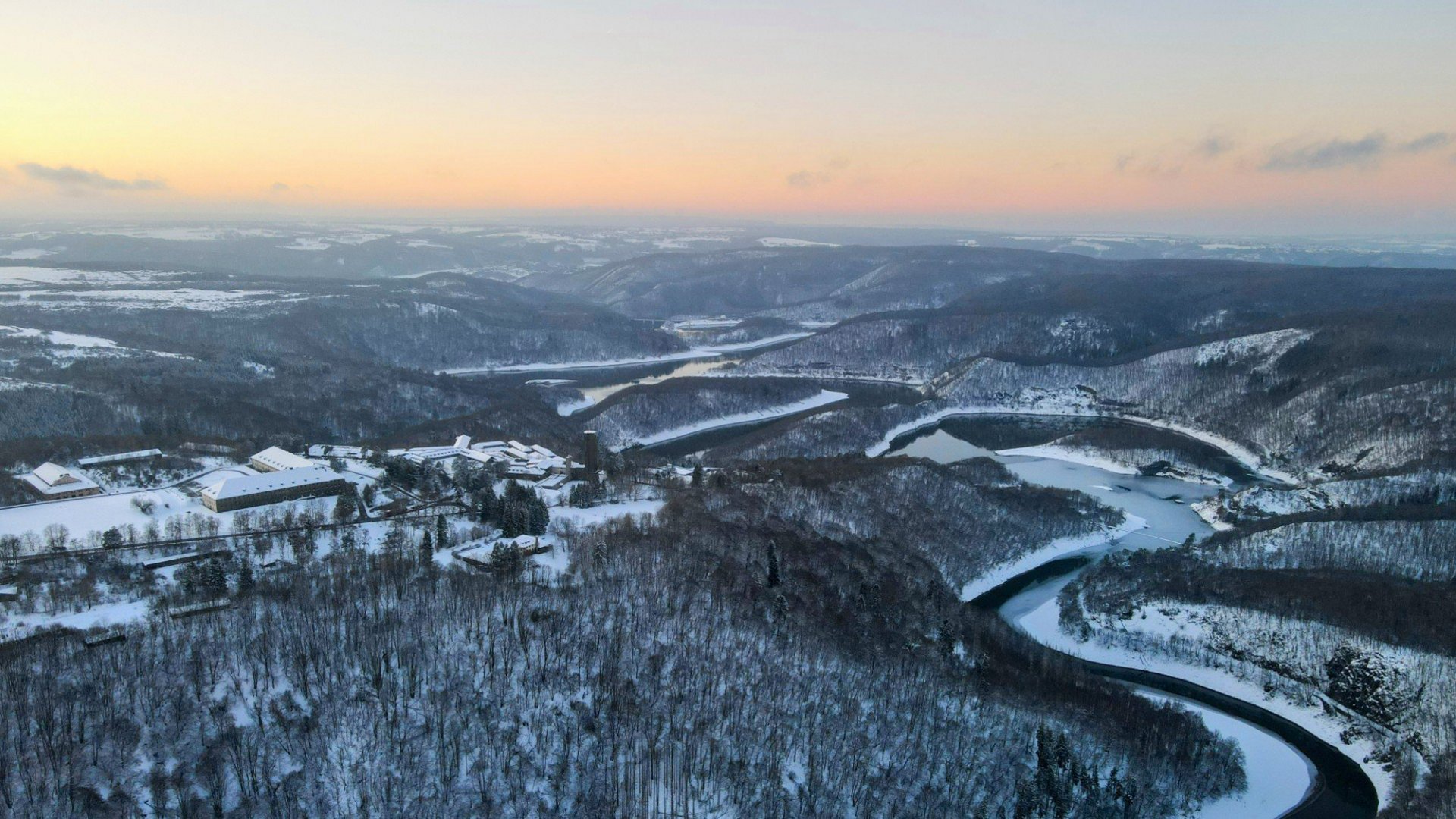 Winterlandschaft mit verschneiten Hügeln, Flusslauf und Gebäuden bei Sonnenuntergang